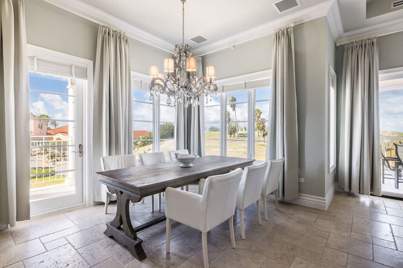 Une salle à manger avec une grande table, des chaises blanches, un lustre et de grandes fenêtres avec des rideaux.