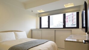 Ein Hotelzimmer mit Bett, Schreibtisch, einem Fenster mit Jalousien und Blick auf Gebäude.