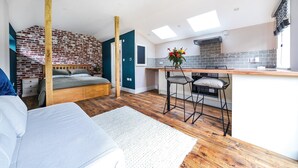 Een moderne slaapkamer met een houten bed, een klein zithoekje met een barkruk en een keuken met een tegelachtergrond.