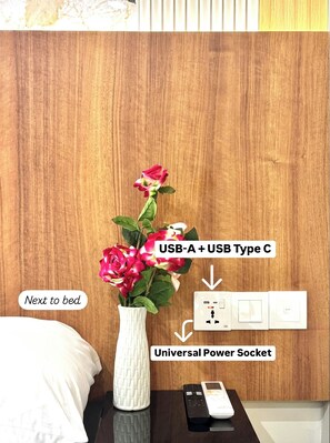 Pared de madera con un jarrón blanco lleno de flores rosadas, un enchufe universal y una cama con almohada blanca.