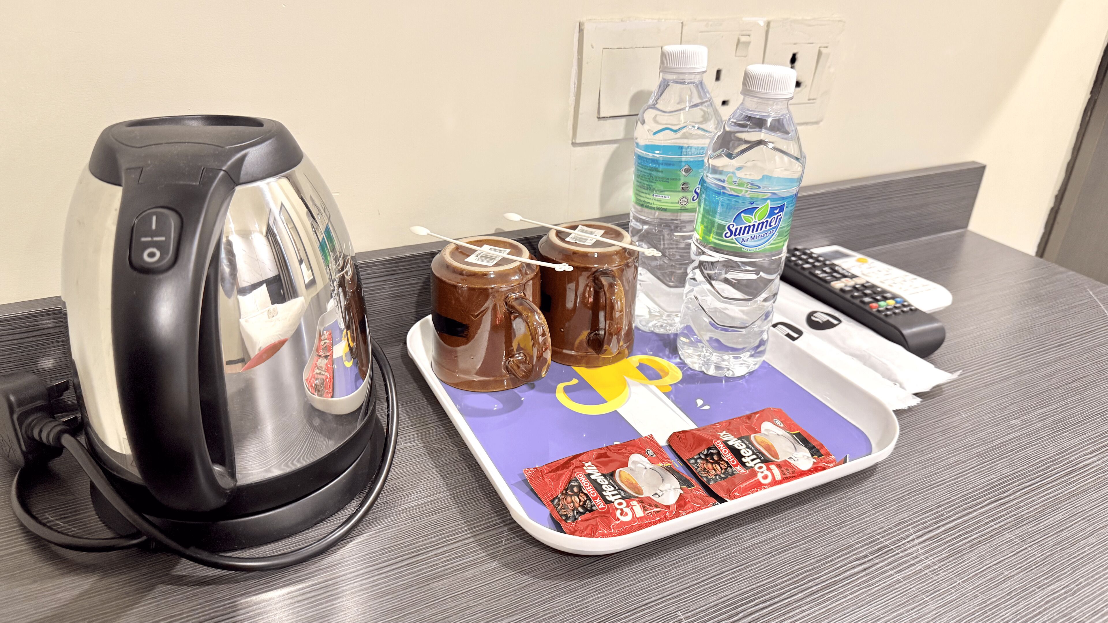 A hotel room with a kettle, two mugs, two bottles of water, a tray with snacks, and a remote control.