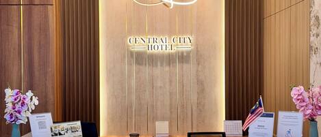 A reception desk with a wooden panel design, a neon sign reading "Central City Hotel", and a flag of Malaysia.