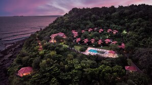 Un resort con varias cabañas de techo rojo rodeadas de abundante vegetación, una piscina y vistas al mar al atardecer.