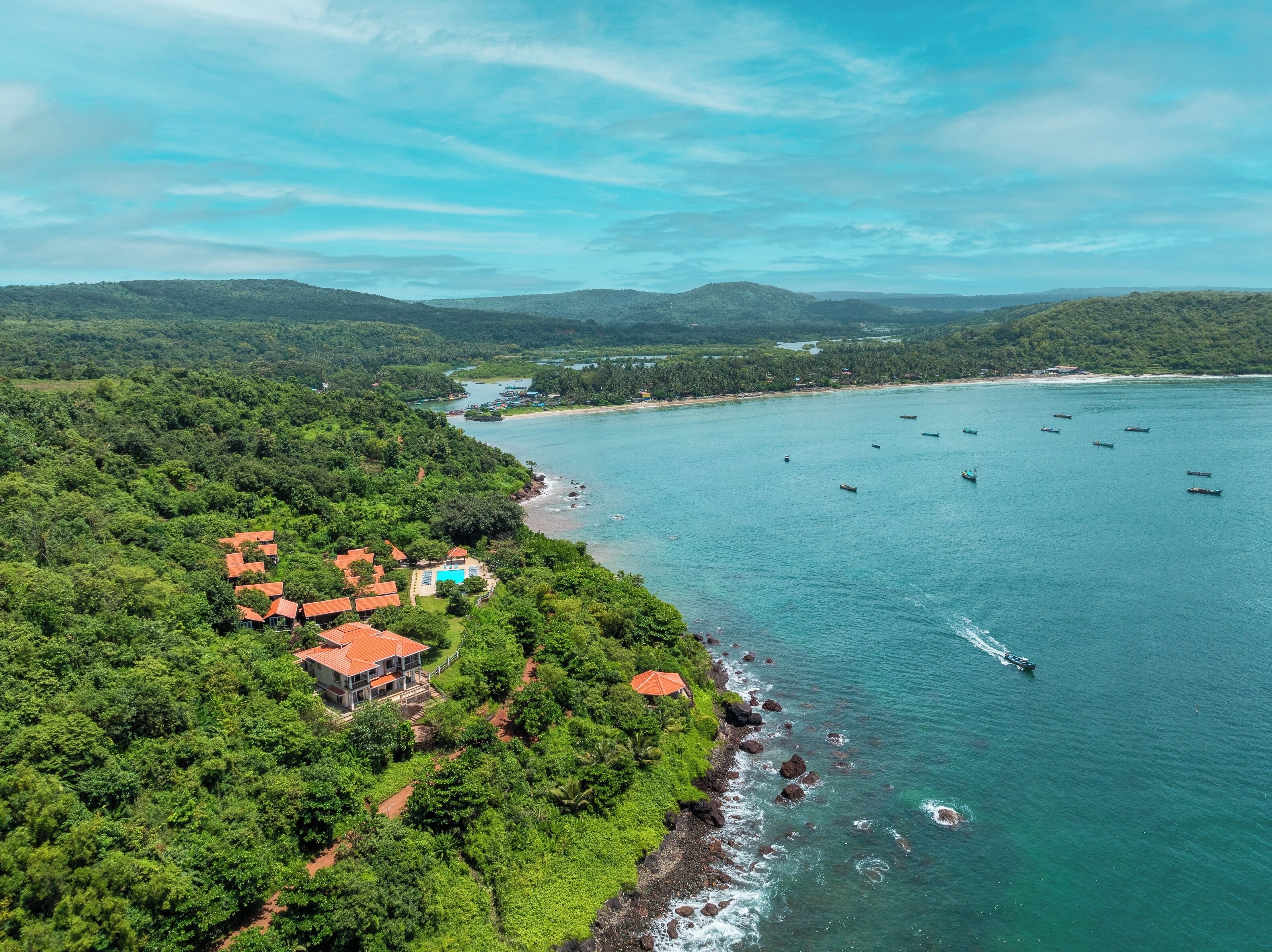 An aerial view of a coastal area with a beach, boats, and a cluster of buildings with red roofs.