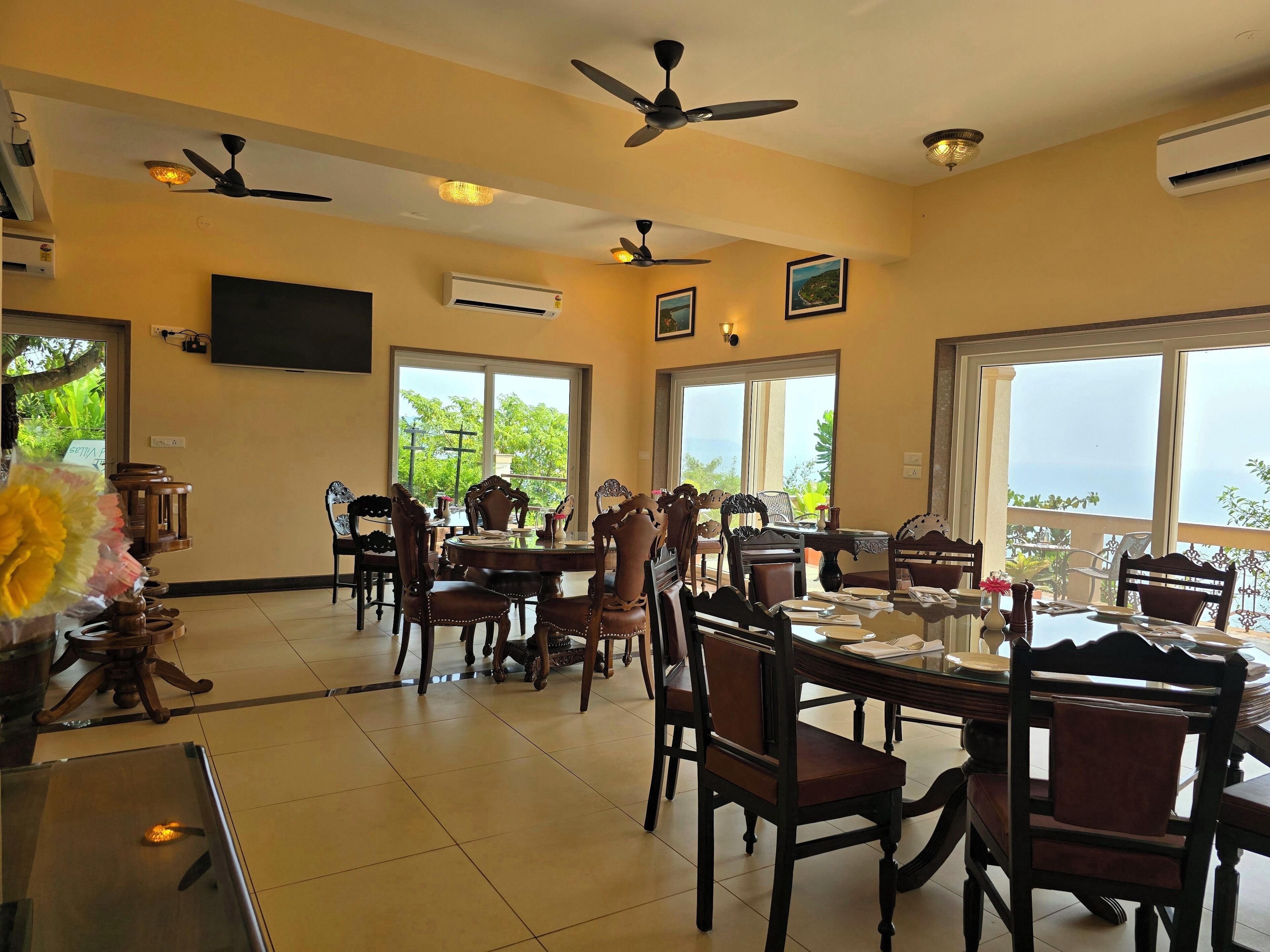A dining area with multiple tables and chairs, ceiling fans, and large windows offering a view of greenery.