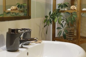 Ein Badezimmer mit einem weißen Waschbecken, einem Chromarmaturen und einem Spiegel darüber. Hinter dem Spiegel befinden sich Pflanzen auf einem Regal.