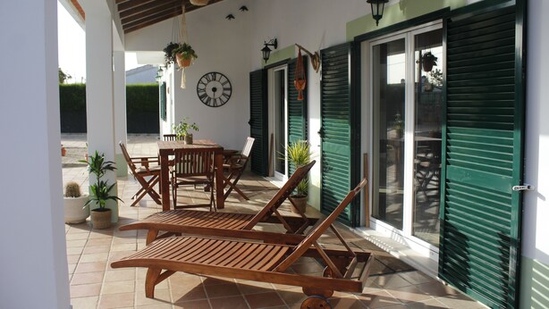 A covered patio with wooden furniture, potted plants, and a wall clock. - Casa Delbon (Aljezur)