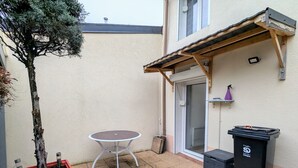 A small patio area with a round table and a potted tree, a trash bin, and a door leading to a covered entrance.