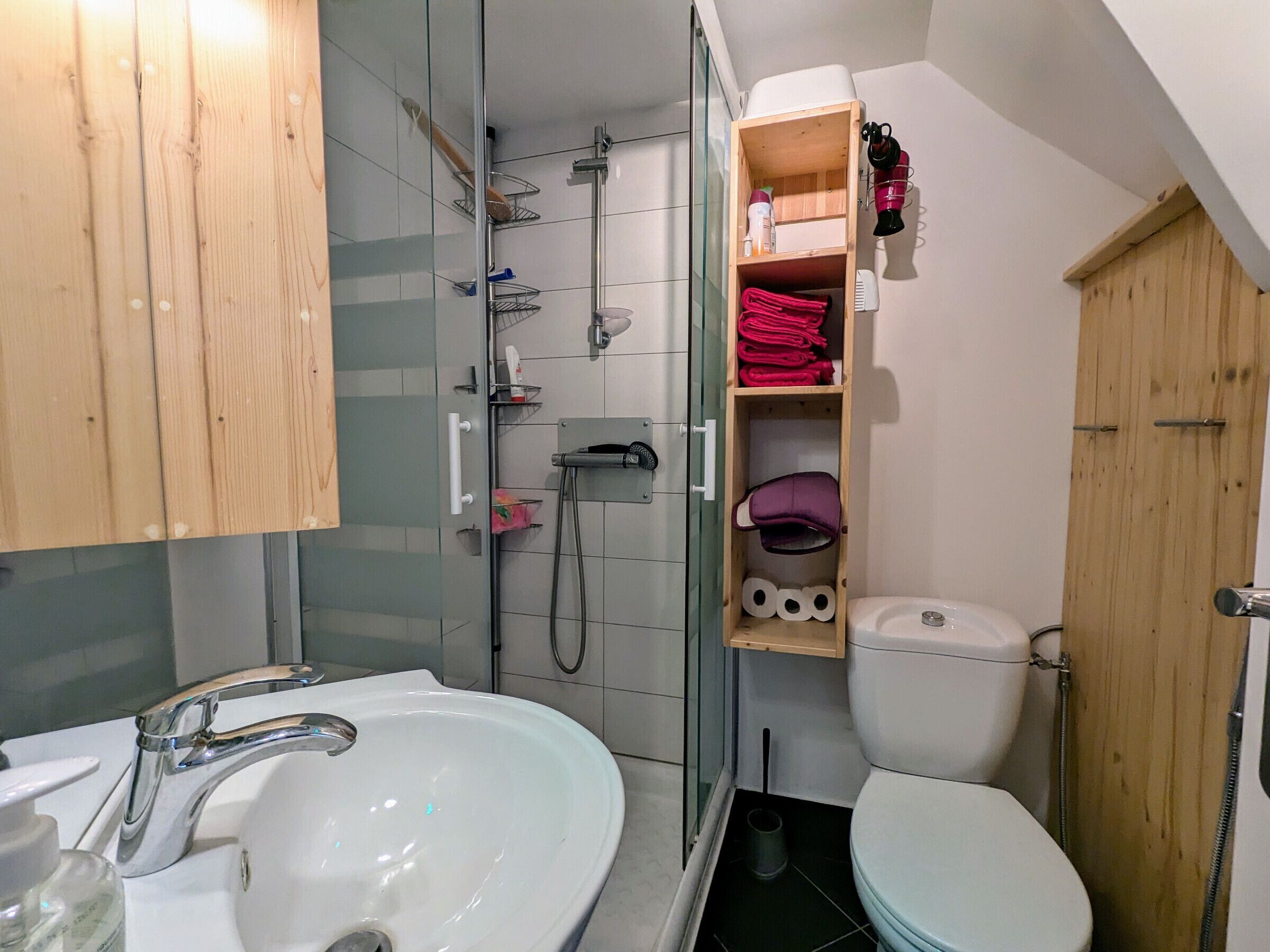 Baño con lavamanos blanco, inodoro y zona de ducha. Hay un estante de madera con toallas y artículos de aseo.