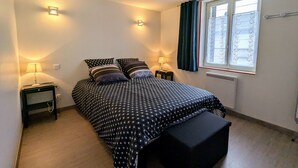 A bedroom with a bed, two bedside tables, a bench, a window with curtains, and wall-mounted lights.
