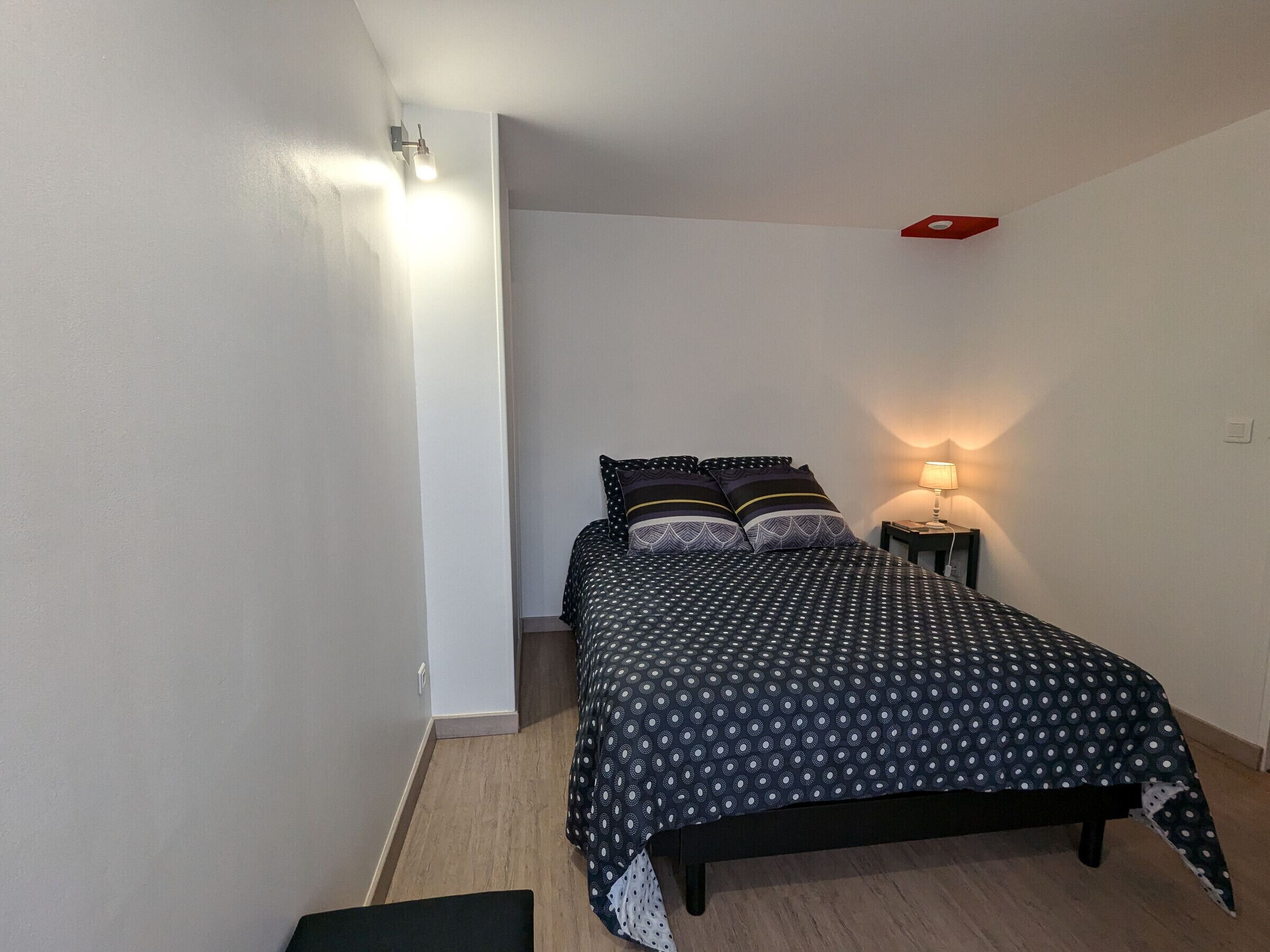 A small, neatly arranged bedroom with a bed, a bedside lamp, and a wall-mounted light fixture.