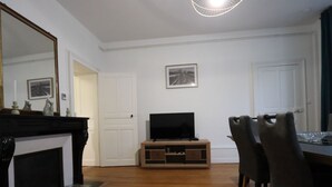 Un salon avec une cheminée, un téléviseur posé sur un meuble en bois et un coin repas avec des chaises.