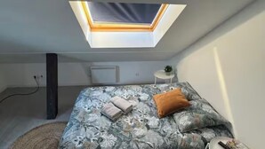 Quarto com claraboia, cama com roupa de cama florida, mesa de cabeceira pequena e tapete redondo.