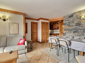 A cozy living room with a grey sofa, a red pillow, and a small wooden table. A dining area with a table set for four is visible, featuring a patterned tablecloth and chairs. The room has wooden doors and a stone wall, with a kitchen area in the background.