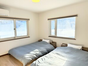 Two single beds with wooden headboards, large windows offering a view of the outdoors, and air conditioning for a comfortable stay.