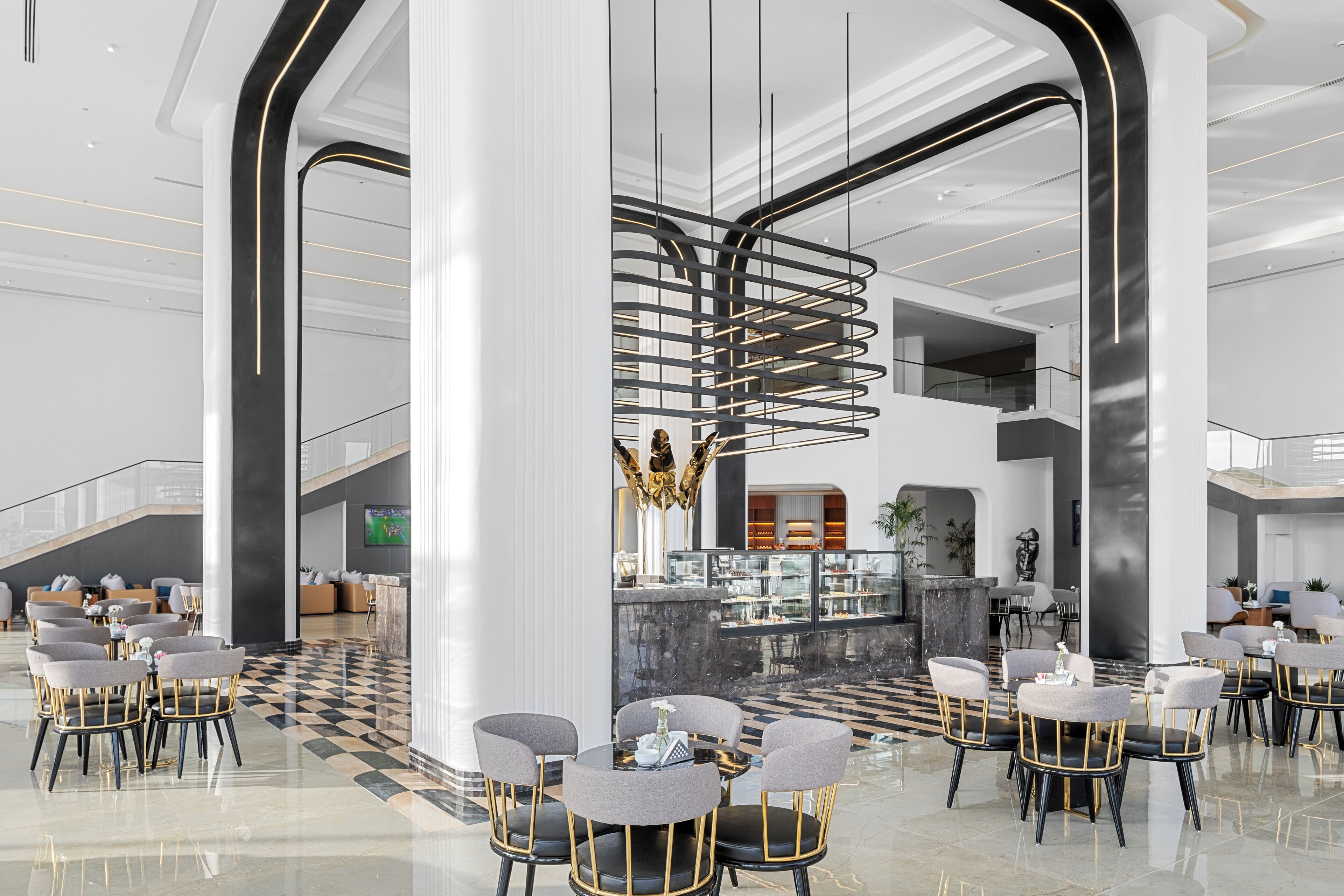 Um hall de hotel moderno com piso xadrez, uma escadaria central e áreas de estar com mesas e cadeiras.