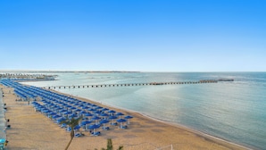Una spiaggia con file di ombrelloni blu e lettini, un molo che si protende nell'acqua, e un cielo sereno.