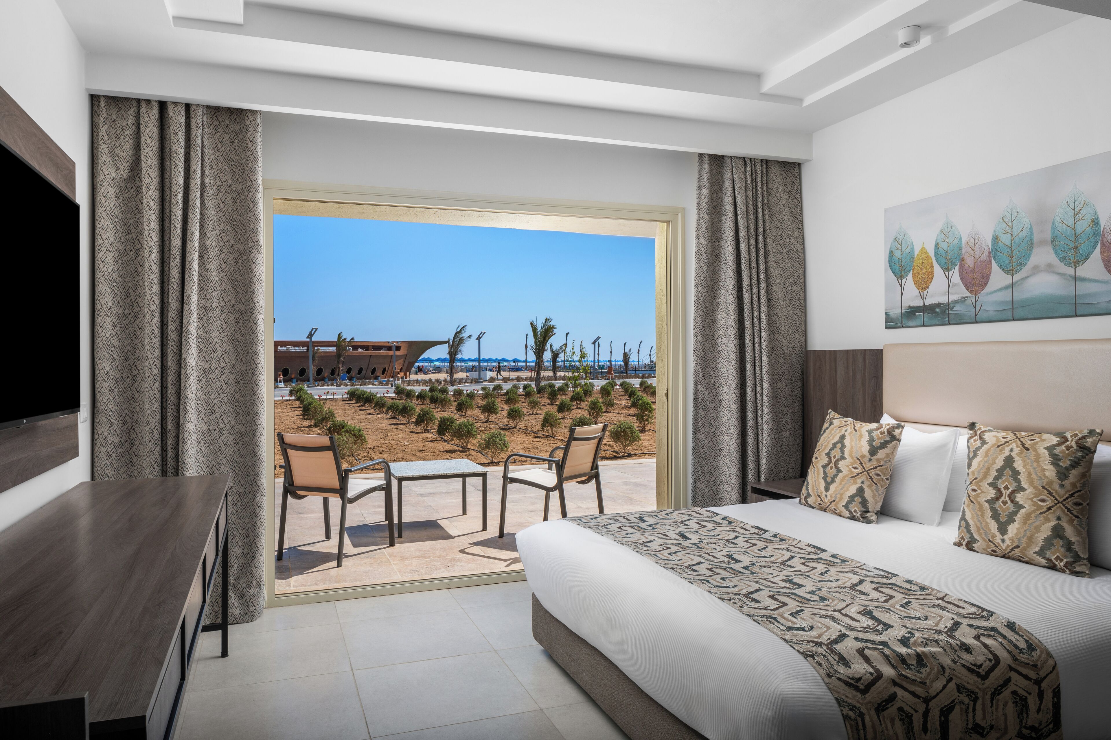 Habitación de hotel con cama, televisión, una mesa con sillas y vista a una playa y palmeras.