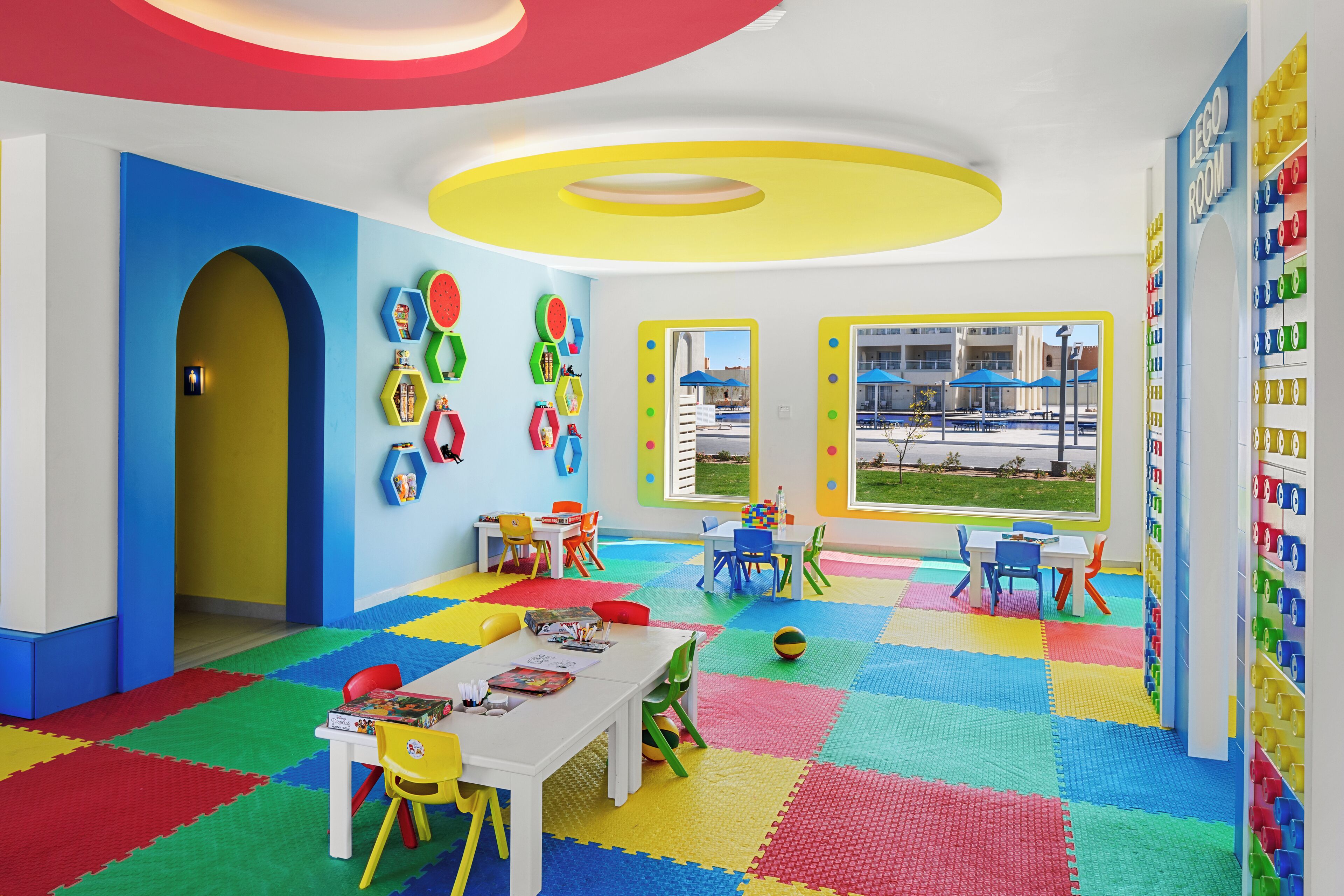 Ein helles Kinderzimmer mit kariertem Boden, Tischen, Stühlen und lehrreichen Wanddekorationen.