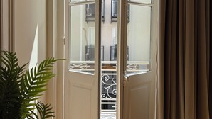 A room with a wooden floor, a potted plant, a door with glass panels, and a view of a balcony with railings. - Generation 6 (Athens)