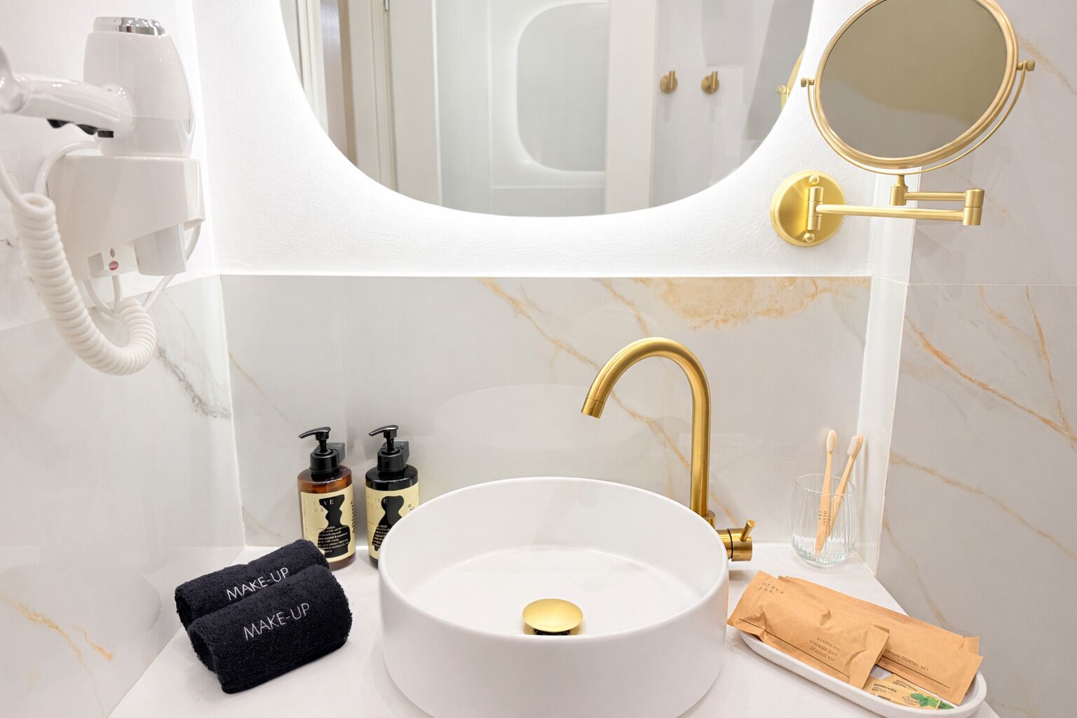 A bathroom with a white sink, gold faucet, and a hairdryer on the wall.