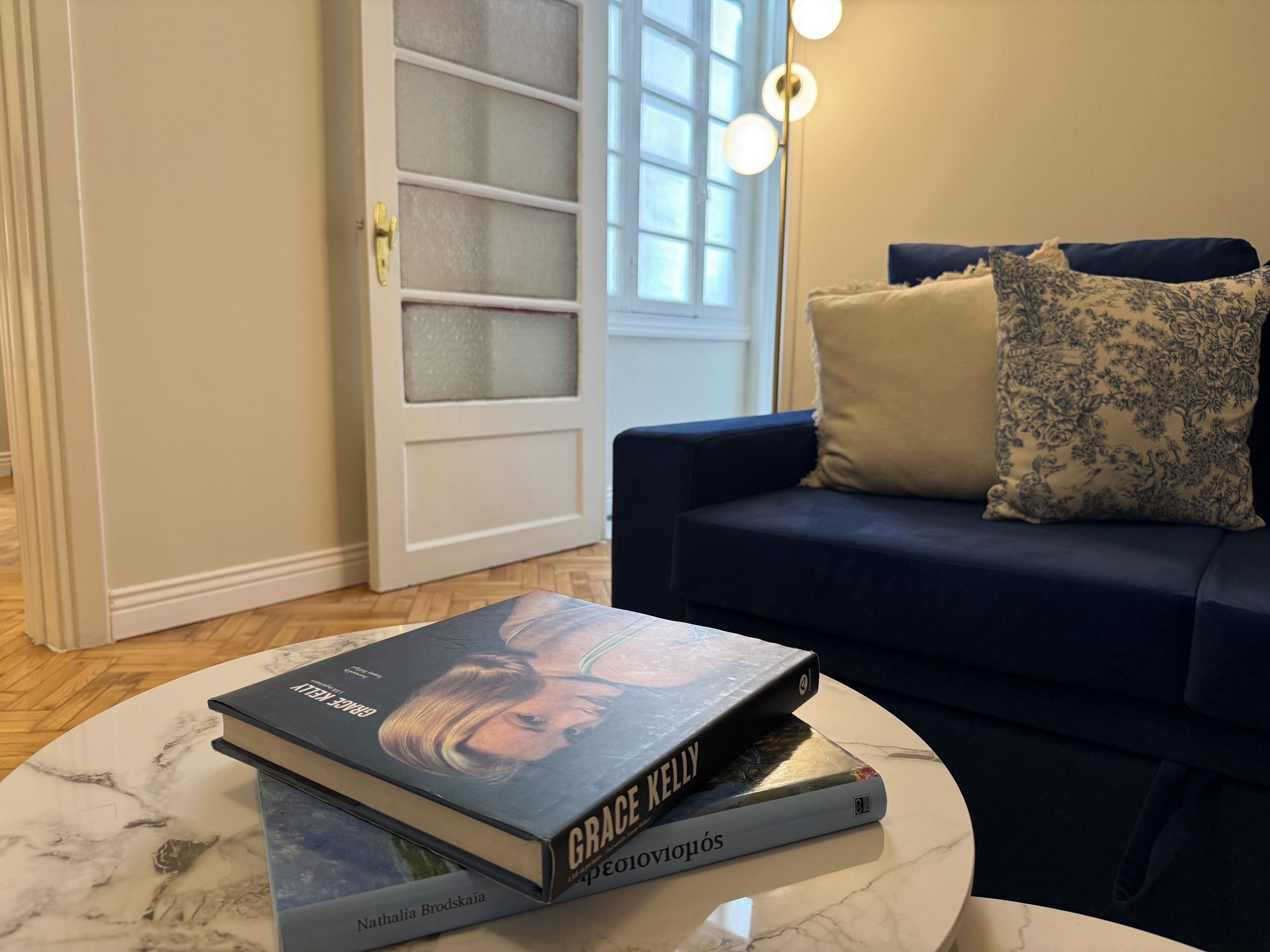 Ein Zimmer mit einem dunkelblauen Sofa, einem Marmor-Couchtisch mit Büchern, einer Tür mit Milchglasfenstern und einer Stehlampe.