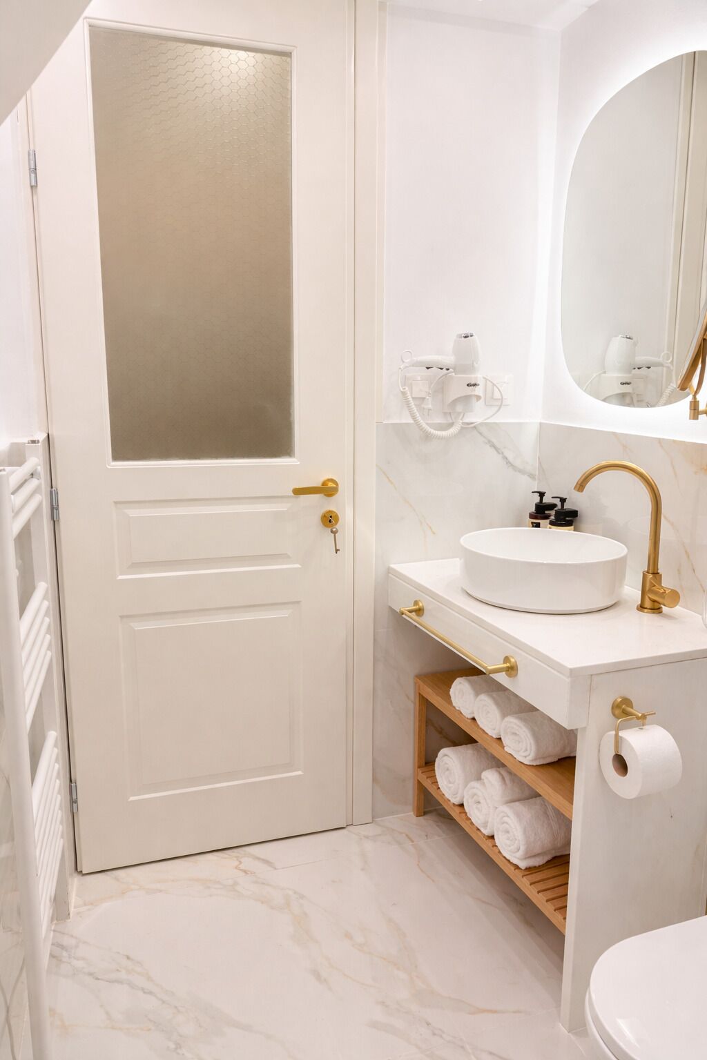 A bathroom with a white door, a white sink with gold fixtures, a mirror, and a towel shelf.