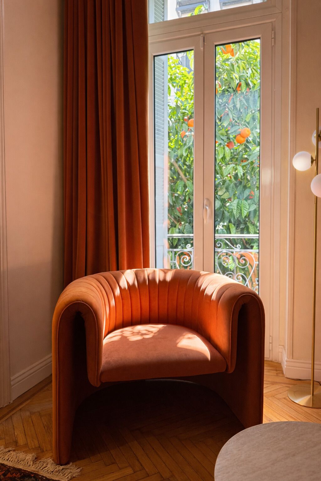 Une pièce avec un fauteuil à dossier incurvé et revêtement en tissu, une grande fenêtre avec des rideaux et une vue sur la verdure à l’extérieur.