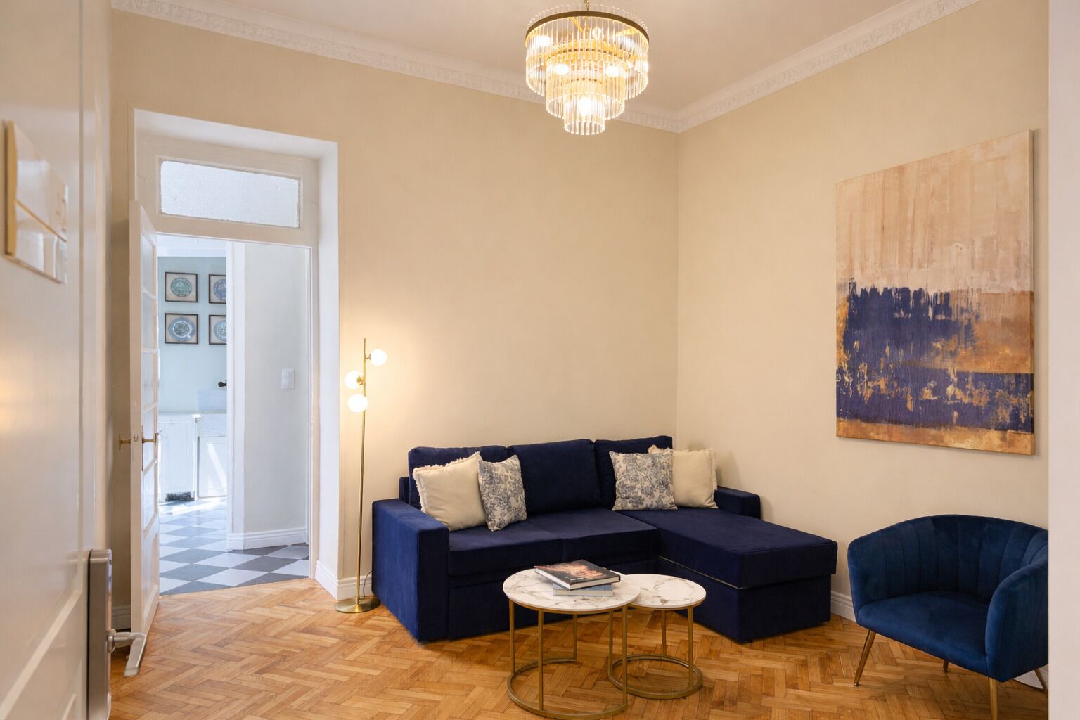 A living room with a dark blue sofa, a round coffee table, a blue armchair, a chandelier, and a painting on the wall.
