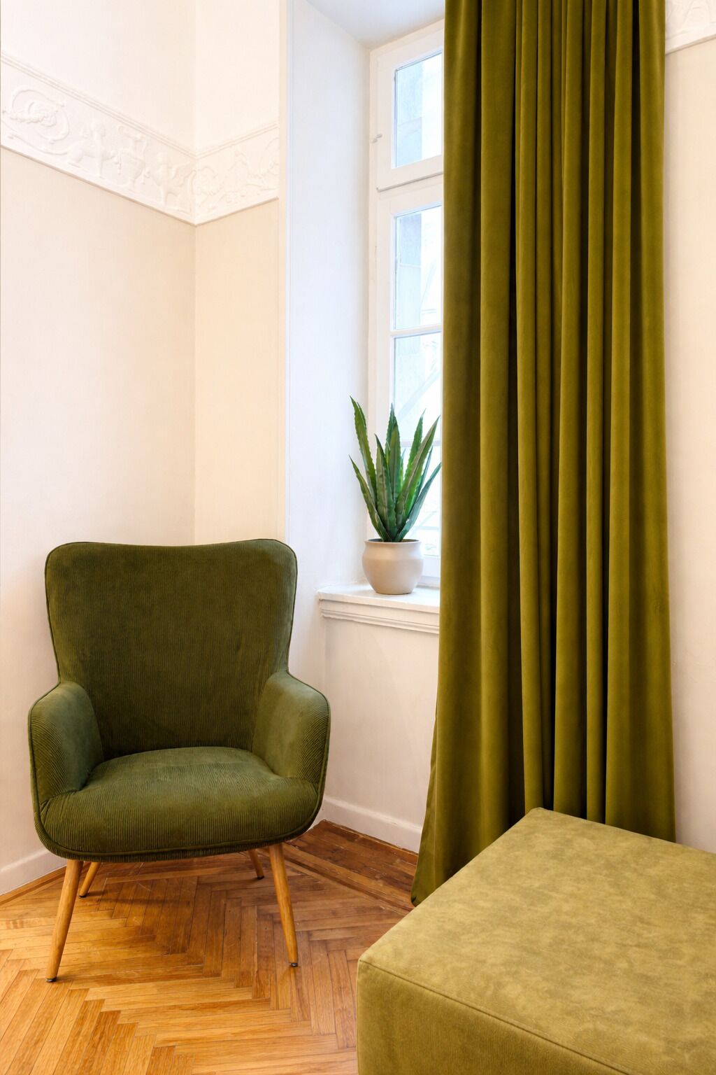 Une pièce avec un fauteuil vert, un pouf vert, une plante en pot et une fenêtre avec des rideaux.