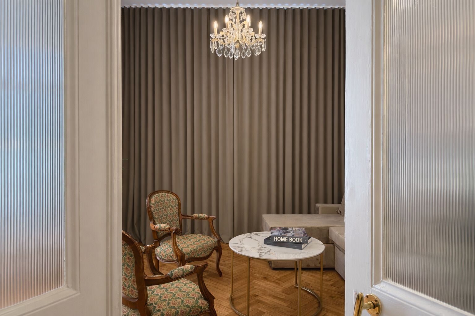 A room with a chandelier, a patterned armchair, a sofa, and a round table with a book.