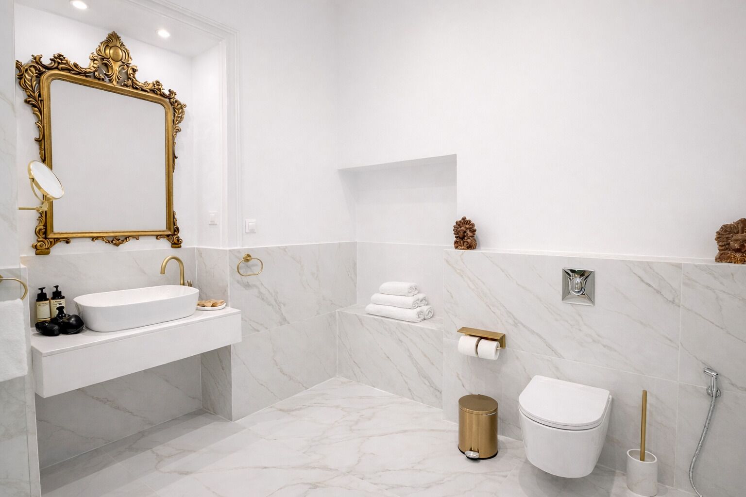 Ein Badezimmer mit weißem Waschbecken, einem spiegeln Spiegel im Goldrahmen, Marmorwänden, einer Toilette und einer Dusche.