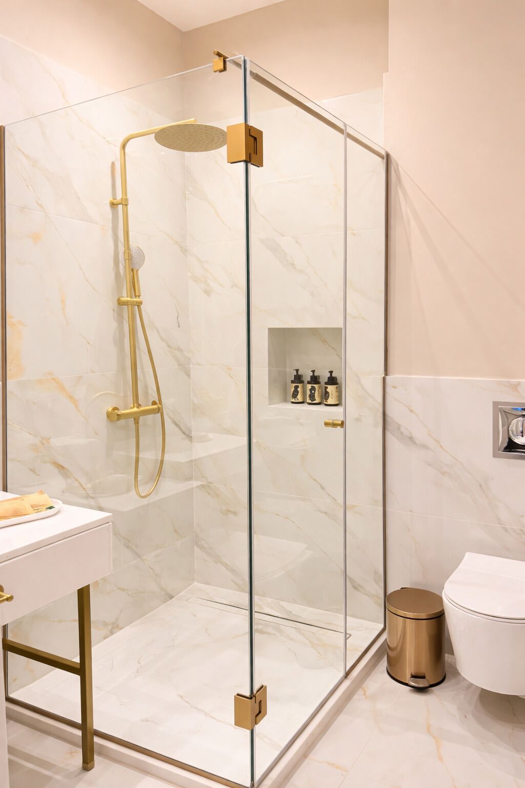 A modern bathroom with a glass-enclosed shower, marble walls, a white sink, and a gold-colored trash bin.