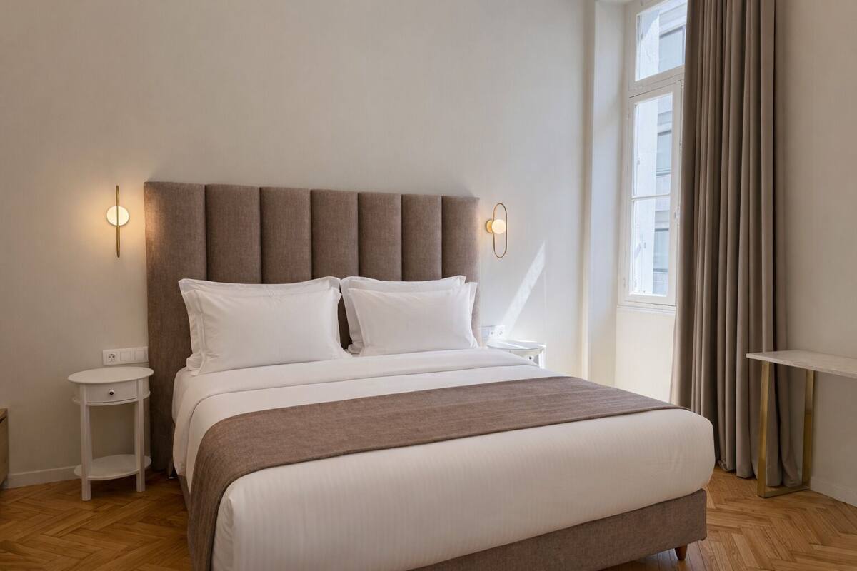 A modern hotel room with a large bed, a bedside table, a nightstand, and a window with curtains.