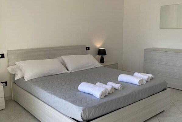 A neatly made bed with a grey quilt, white pillows, and rolled towels on the side.
