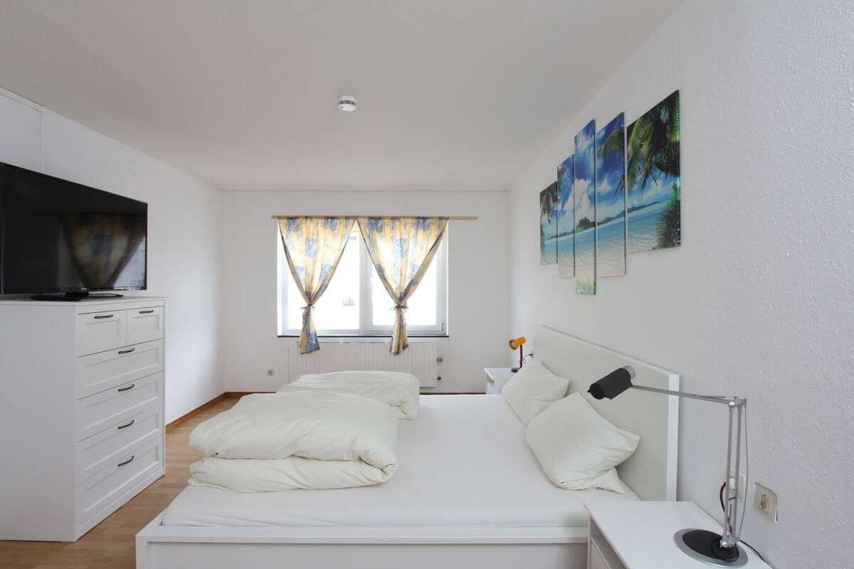 A bedroom with a bed, a TV, a desk, and a window with curtains.