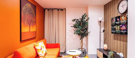 Una sala de estar moderna con un sofá naranja intenso, un tapete geométrico y colorido, una mesita redonda pequeña, una planta en maceta y un televisor de pared que muestra carteles de películas.