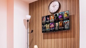 A modern living room with a wooden panel wall, a large clock, and a TV mounted on the wall displaying various movie posters. - Cosy Bradford Apartment With City Views (Bradford)