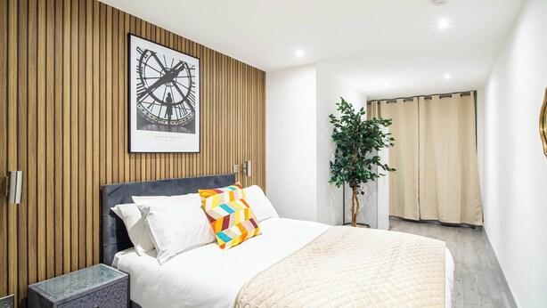 A modern bedroom with a bed, a nightstand, a wall-mounted clock, a potted plant, and a view of the bathroom. - Cosy Bradford Apartment With City Views (Bradford)