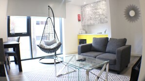 A modern living room with a glass coffee table, a grey sofa, a hanging chair, and a large window offering a view of the outside.