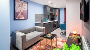 A modern living room with a grey sofa, a small black table with a gold frame, and a pink rug. There is a kitchen in the background with grey cabinets and a blue chair. - Bradford City Apartment - Bolton Road Oasis (Bradford)