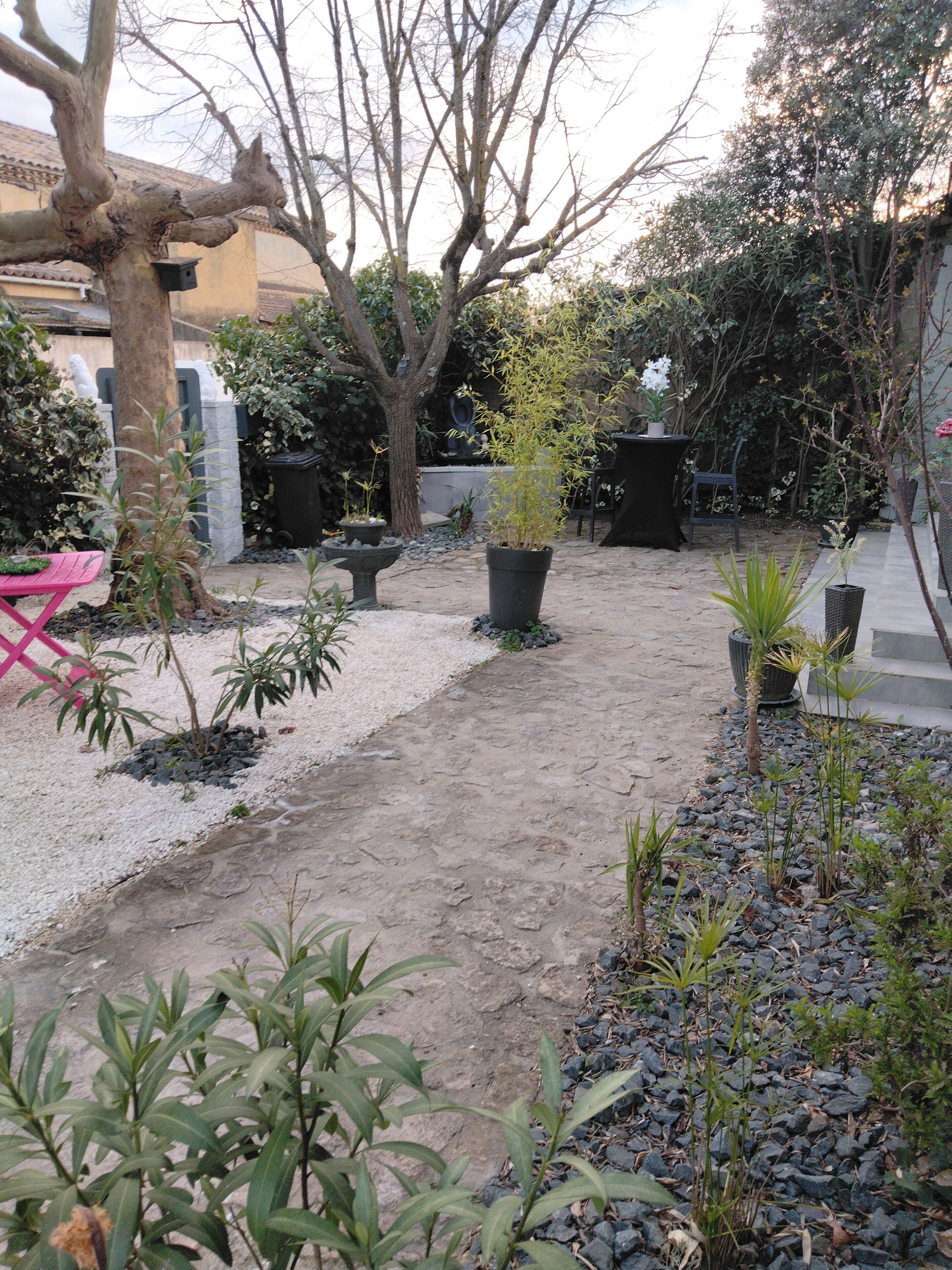 Un jardin avec un chemin de gravier, des plantes en pot, une chaise rose et un arbre.