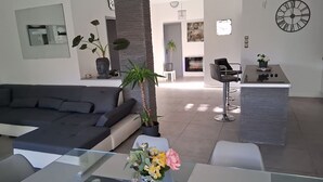Una sala moderna con un sofá, una mesa de centro y una planta en maceta. Se aprecia una cocina con una barra y una mesa de comedor al fondo.