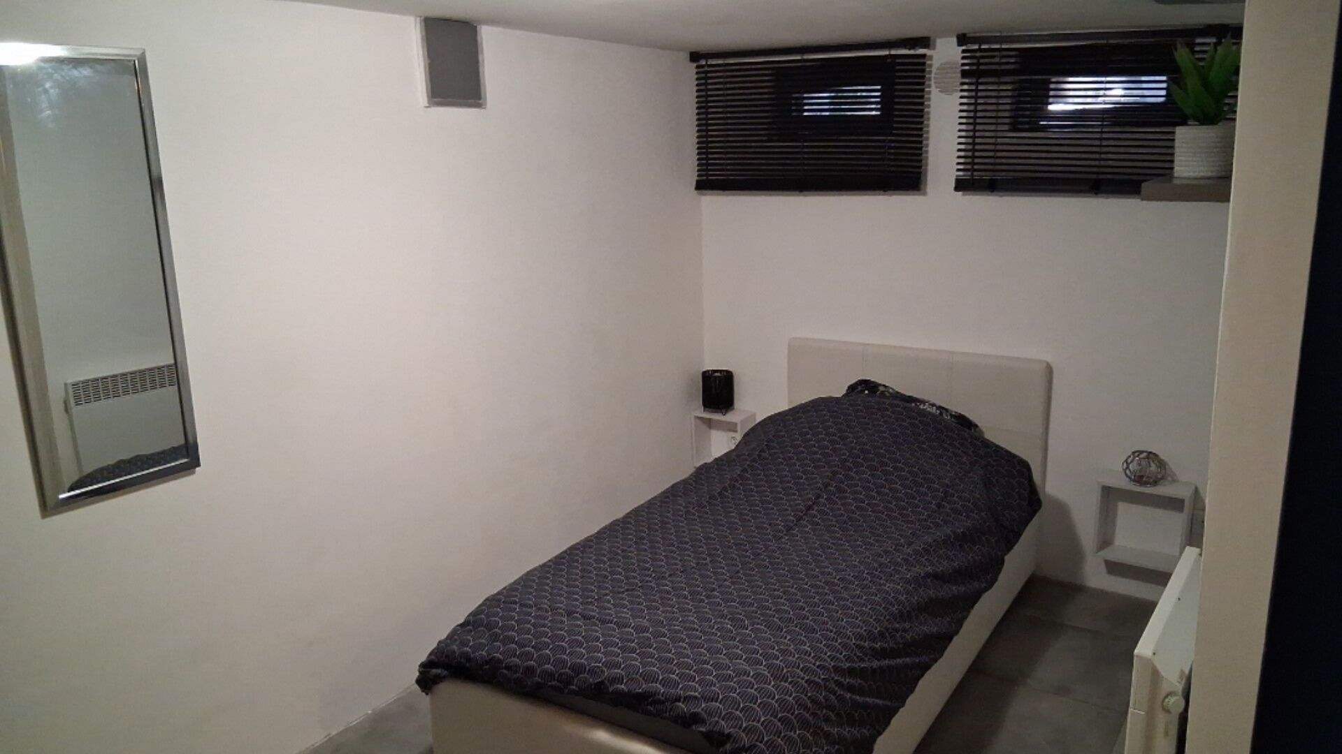 Ein kleines Zimmer mit einem Einzelbett, einer schlichten weißen Wand, einem Nachttisch mit Lampe und einem Fenster mit Jalousien.