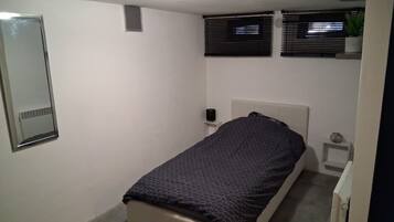 Ein kleines Zimmer mit einem Einzelbett, einer schlichten weißen Wand, einem Nachttisch mit Lampe und einem Fenster mit Jalousien.
