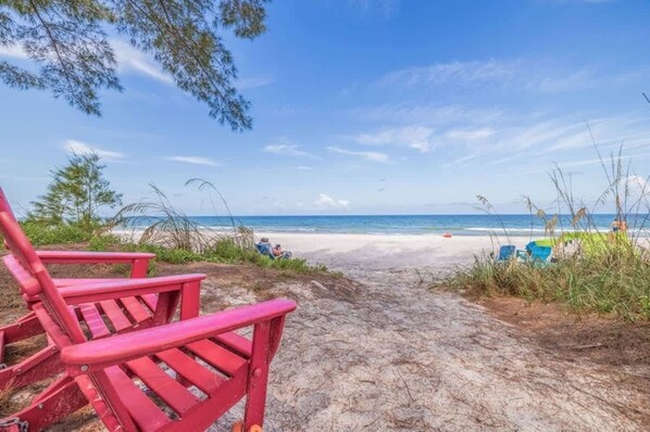 Un sentier bordant la plage, avec des chaises rouges qui mènent au rivage, un ciel dégagé et une mer calme.