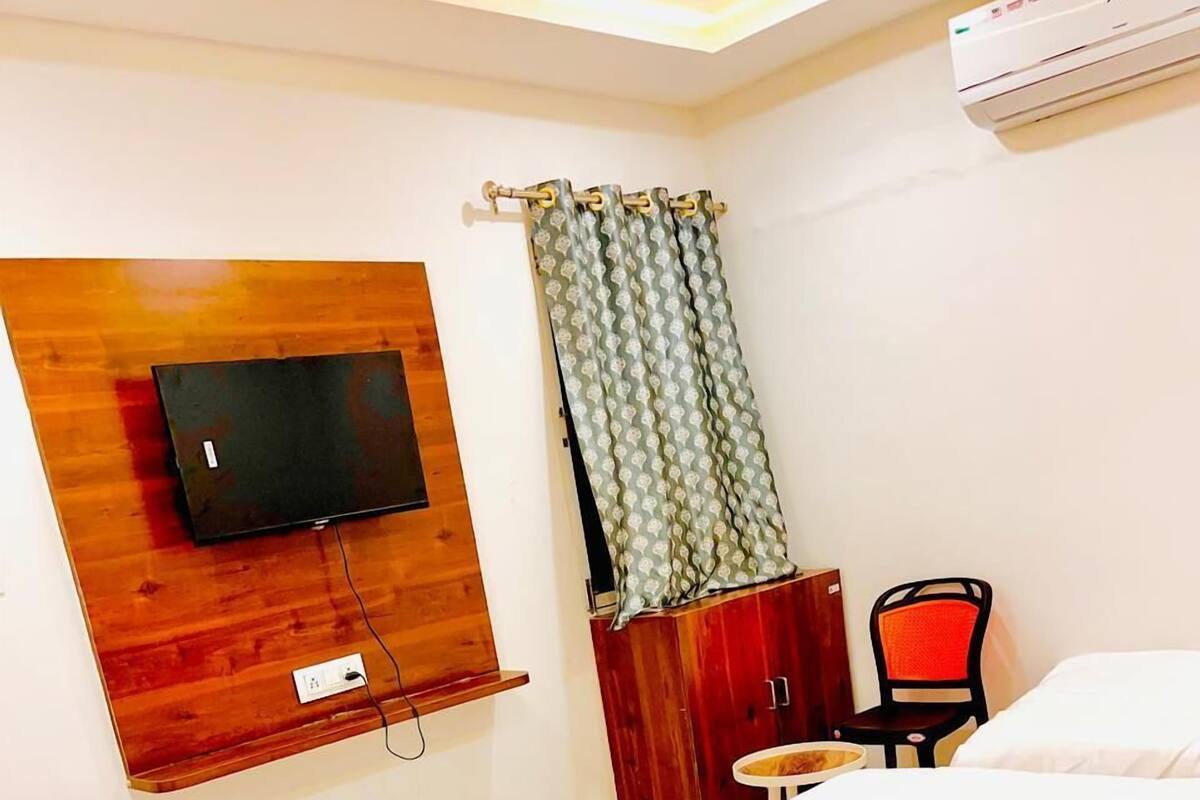 A hotel room with a bed, a TV mounted on a wooden panel, a wardrobe, a chair, and an air conditioning unit.