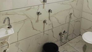 A bathroom with white marble tiles, a showerhead, a sink, a toilet, and a black bucket. - Hotel Shree Kedar Palace (Ujjain)