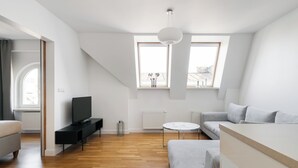 Un soggiorno moderno con un divano, una TV e un tavolo rotondo. Due finestre offrono luce naturale e vista sulla città.