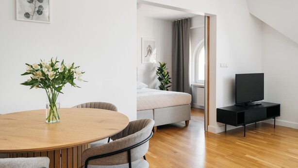Ein modernes Wohnzimmer mit einem runden Holztisch, zwei grau gepolsterten Sessel, einer schwarzen TV-Konsole, einem Bett mit grauer Bettwäsche und einem Fenster mit Vorhängen.